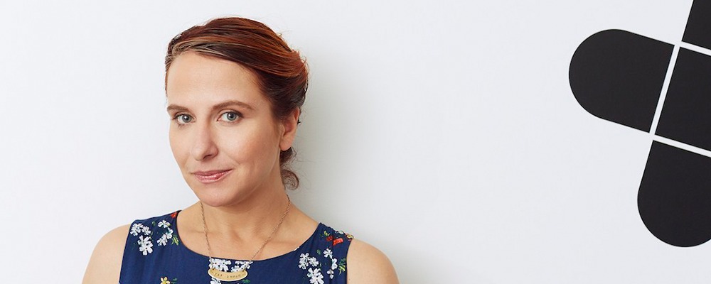 Colour image of Anna Pickard's head and shoulders. She is standing in front of a white wall. Her hair is dark red. Her sleeveless dress is blue with small white flowers. She wears a gold necklace.