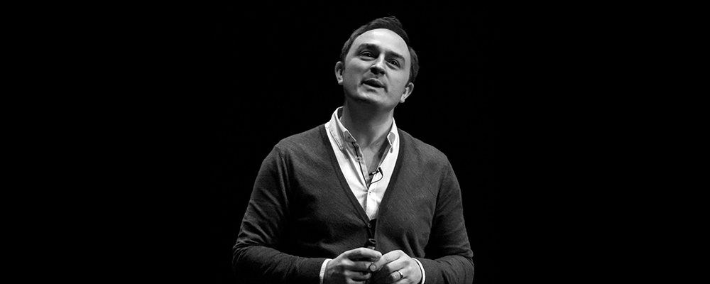 Black and white image of Tom Cheesewright's head a shoulders. He is standing in front of a black background. He wears an open-necked white shirt and a cardigan.