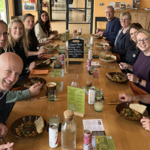 A group shot of the mark-making* team sitting at a long wooden table inside the FarmEd café, smiling at the camera while enjoying their meal together.