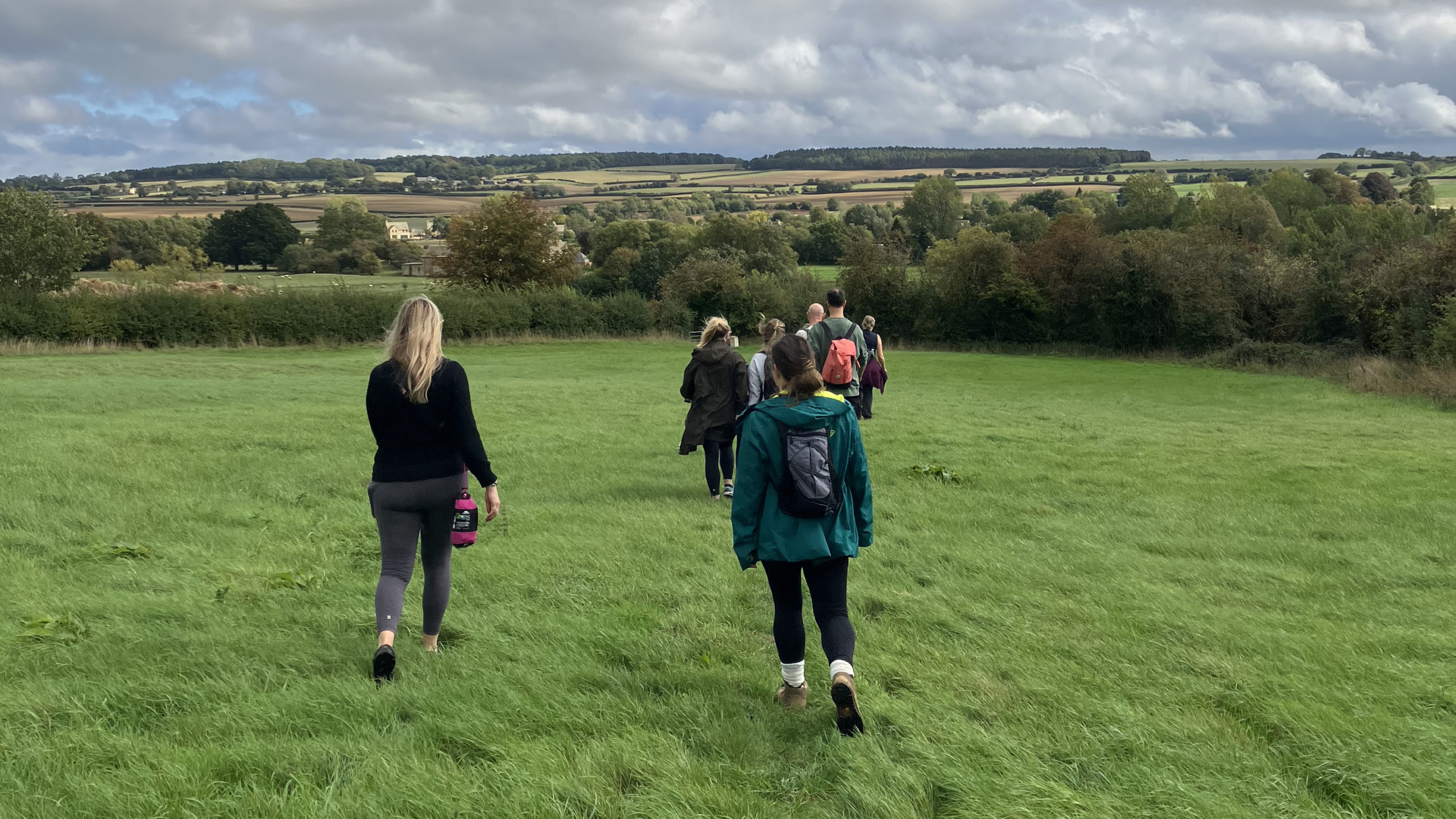 The mark- makers, seen from behind, walking in single file across a large, vibrant green field. In the distance, beyond a line of trees, are the rolling hills and picturesque valleys of the Cotswolds countryside.