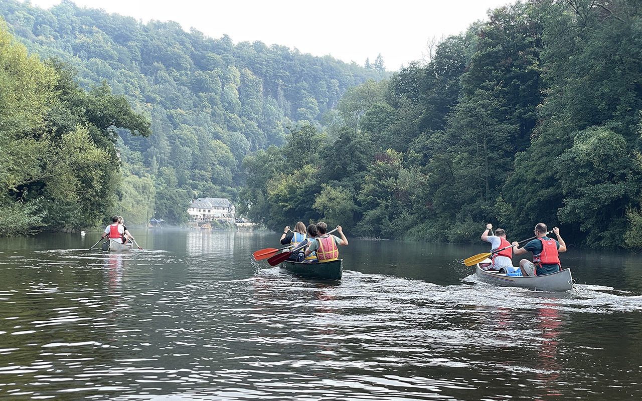 Three canoes with mark-making* team members paddling on calm river surrounded by wooded hills at Symonds Yat