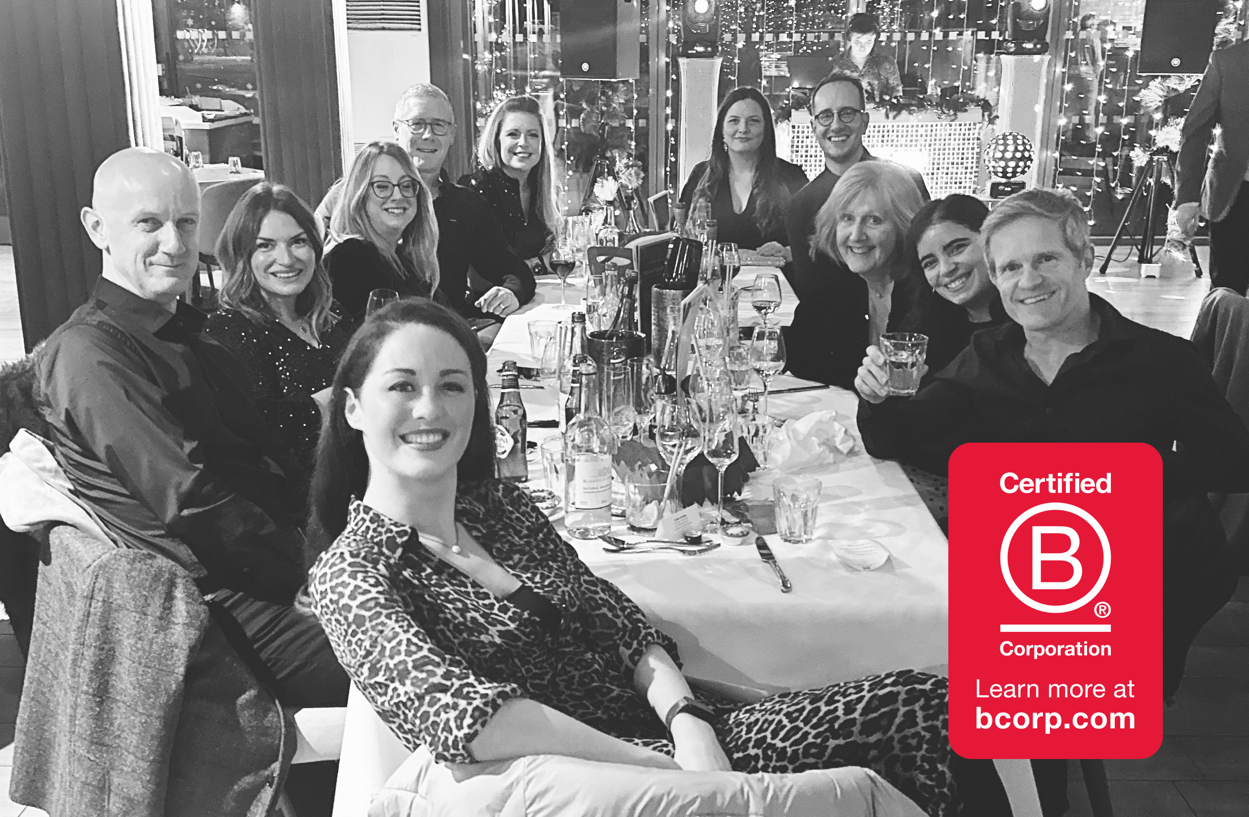 A black and white photograph of the mark-making* team seated together at a restaurant table during their Christmas party, smiling and in good spirits. The Certified B Corporation logo appears in the bottom right of the image.
